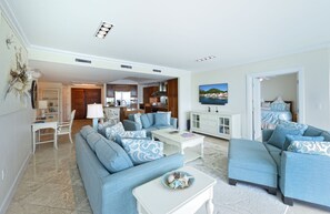 A spacious living room with a large sofa, a coffee table, and a television. There is a kitchen area visible in the background.