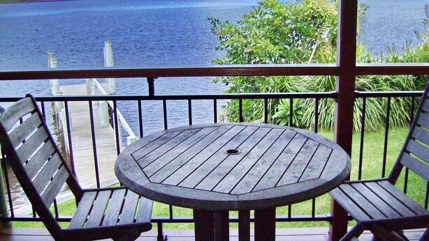 A wooden table and two chairs on a balcony with a view of the water. - Coast & Country Cabin 2 (RAINBOW REACH)