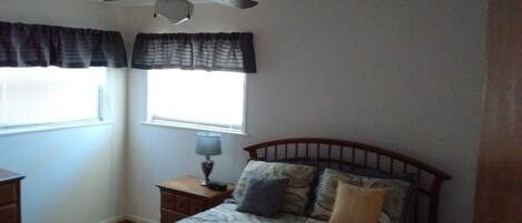 A bedroom with a bed, a nightstand, a lamp, a ceiling fan, and two windows with curtains.