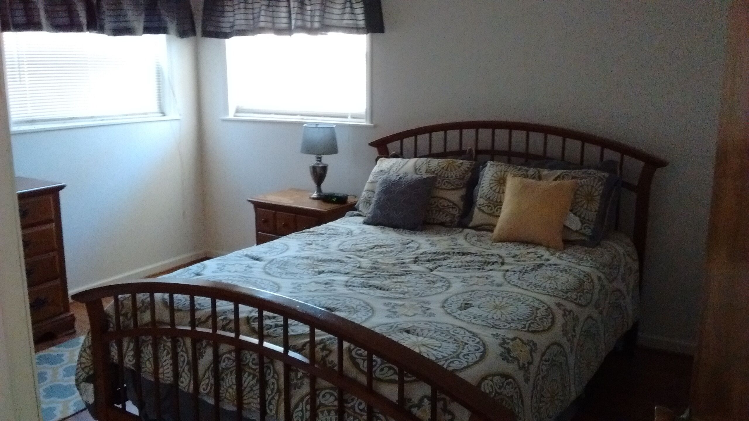 A bedroom with a wooden bed frame, patterned bedding, a bedside table with a lamp, and a window with curtains.
