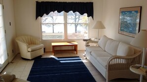 Una sala de estar con un sofá blanco, un sillón blanco, una mesa de centro de madera, un tapete azul, una silla de mimbre, una lámpara y un cuadro enmarcado en la pared.