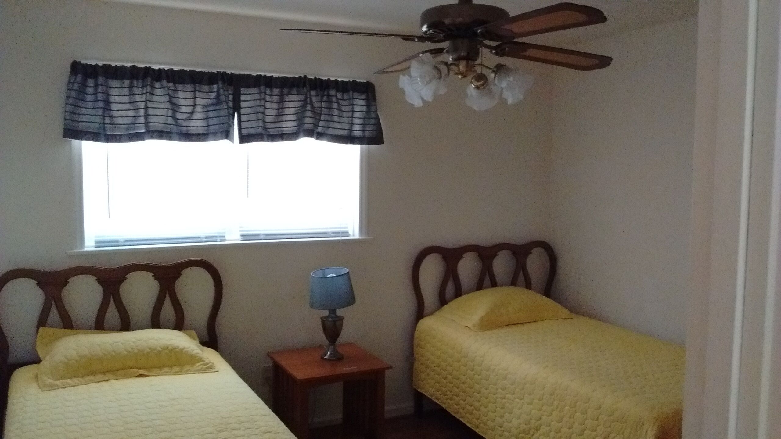 A bedroom with two beds, a window with curtains, a small table with a lamp, and a ceiling fan.