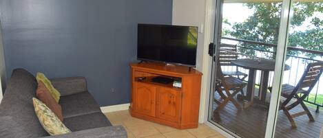 A living room with a grey sofa, a wooden entertainment unit with a flat-screen TV, and a sliding glass door leading to an outdoor patio with wooden furniture.