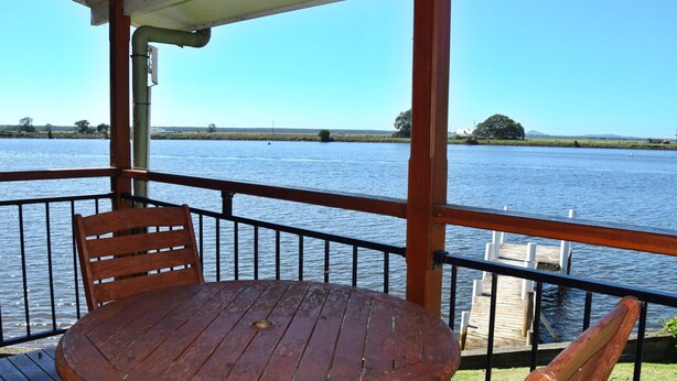 A wooden table and chair on a balcony overlooking a body of water with a clear sky. - Coast & Country Cabin 4 (Rainbow Reach)