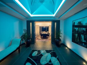 Ein modernes Interieur mit einer blauen Deckenbeleuchtung, einem Essbereich mit Tisch und Stühlen sowie einem großen abstrakten Gemälde an der Wand.