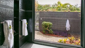 Un baño moderno con paredes de baldosas oscuras, cabina de ducha de vidrio y vistas a un jardín ajardinado.