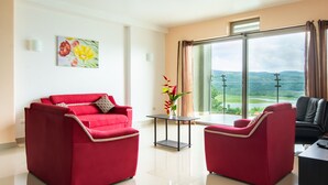 Uma sala de estar moderna com um sofá vermelho, uma poltrona de couro preto e uma mesinha com um vaso de flores. Uma grande janela oferece vista para um lago e colinas.