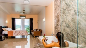Quarto de hotel moderno com cama, sofá, banheira de vidro e vista para o exterior.