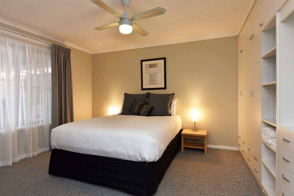 Une chambre à coucher comprenant un lit, une table de chevet, une commode, un ventilateur de plafond et un placard intégré.