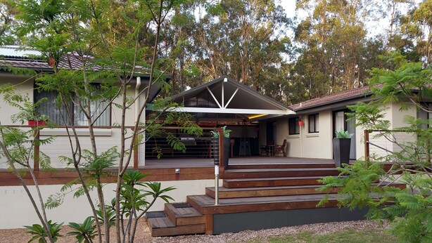 Uma casa moderna com área externa coberta, escadas de madeira e vegetação ao redor.