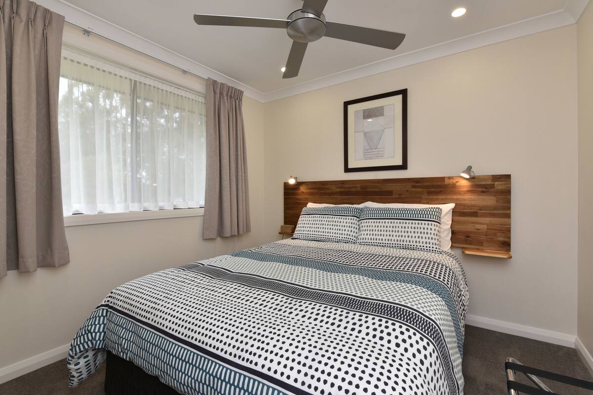 Une chambre à coucher avec un lit, un ventilateur de plafond, une fenêtre avec des rideaux, un tableau encadré au mur et une table de chevet avec une lampe.