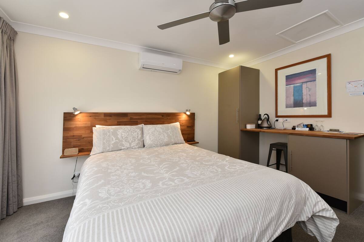 Une chambre à coucher comprenant un lit, un bureau, une chaise, un climatiseur, un ventilateur de plafond et un tableau encadré accroché au mur.