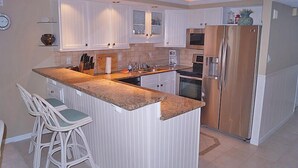 A modern kitchen with white cabinetry, a central island with bar stools, stainless steel appliances, and a clock on the wall. - Ocean Hideaway (Ocean City)