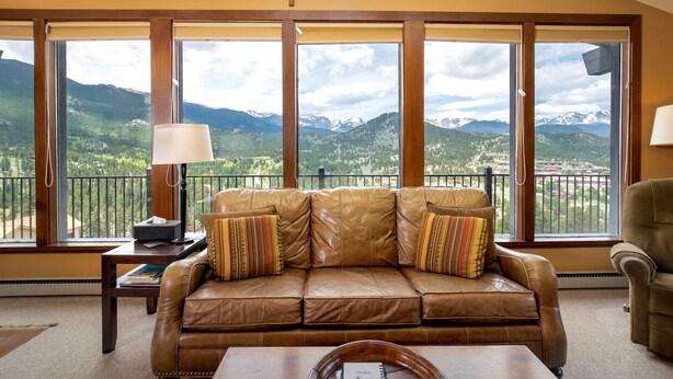 A room with a leather sofa, a table, and a view of mountains through large windows. - Kintzle Family Vacation At Windcliff 4 Bedroom Home by RedAwning (Estes Park)