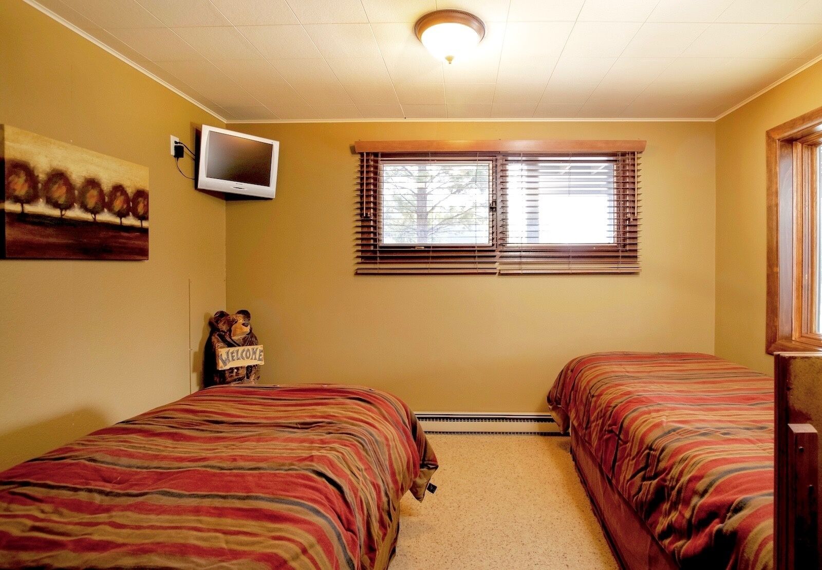 Ein Zimmer mit zwei Betten, einem an der Wand befestigten Fernseher, einem Fenster mit Jalousien und einem Teddybären mit einem 'Welcome'-Schild.