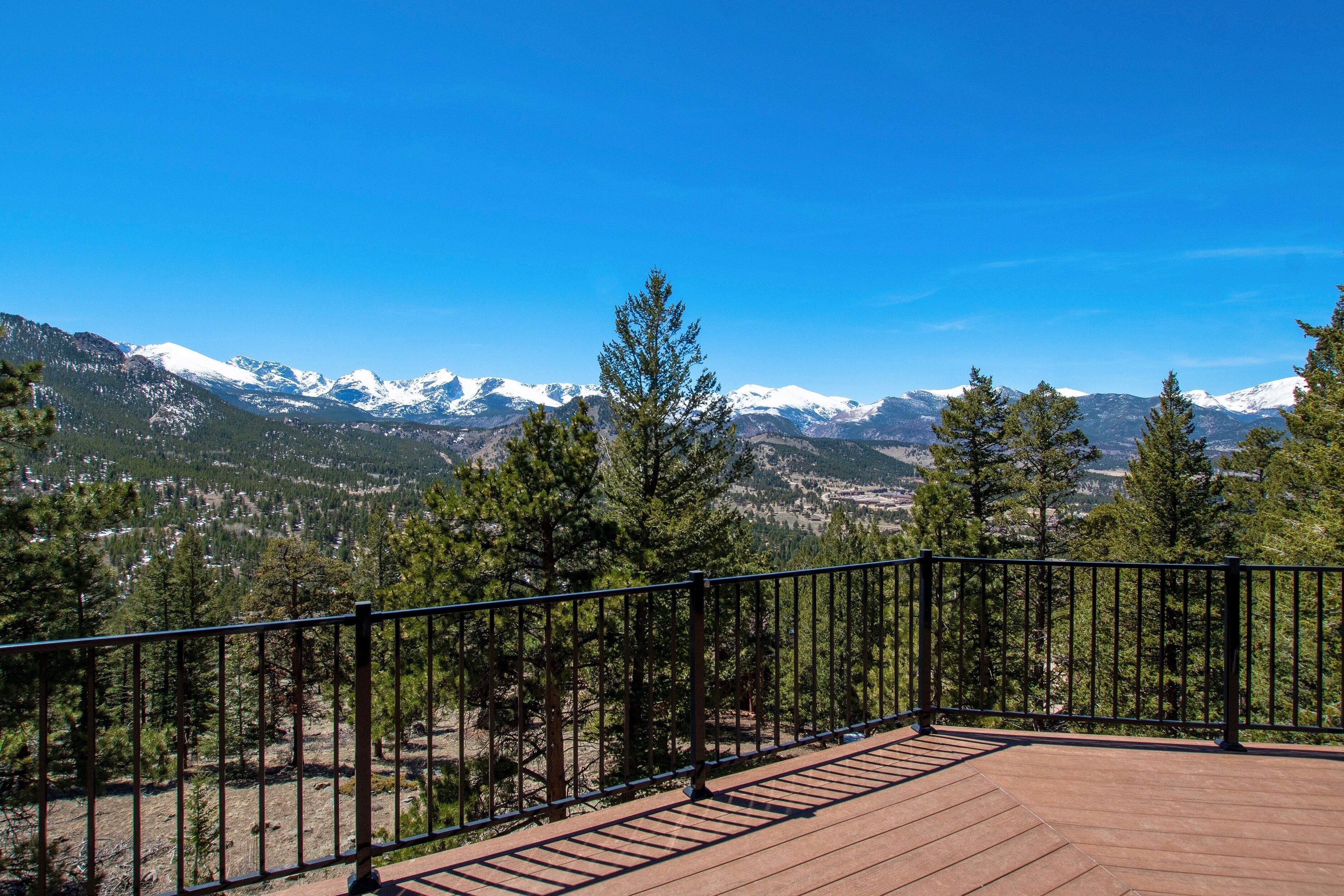 A wooden deck with a metal railing overlooking a mountainous landscape with snow-capped peaks and dense forest.