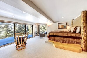 A modern bedroom with a large bed, a wooden chair, and a view of trees through large windows.