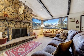 A cozy living room with a stone fireplace, a large window with a mountain view, a leather sofa, and a red patterned rug.