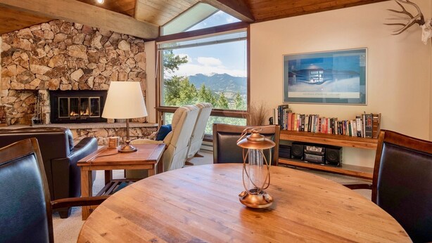 Ein gemütliches Zimmer mit Kamin aus Stein, einem runden Holztisch, Ledersesseln, einem Bücherregal und einem großen Fenster mit Blick auf die Berge.