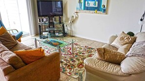 A living room with a glass coffee table, beige sofas, a flat-screen TV, and a decorative rug. - Thunder Island 128c 2 Bedroom Condo by RedAwning (Ocean City)