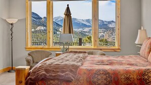 Un dormitorio con un ventanal grande que ofrece vistas a la montaña, una cama con ropa de cama estampada, una silla, una mesita y una lámpara de pie.