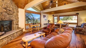 A spacious living room with a stone fireplace, leather sofa, wooden coffee table, and large windows offering a view of the outdoors.