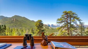 A wooden table with binoculars, a notebook, and a pen, overlooking a mountainous landscape with trees and a cabin.