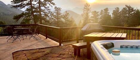A wooden deck with a hot tub, outdoor furniture, and a view of mountains during sunset.