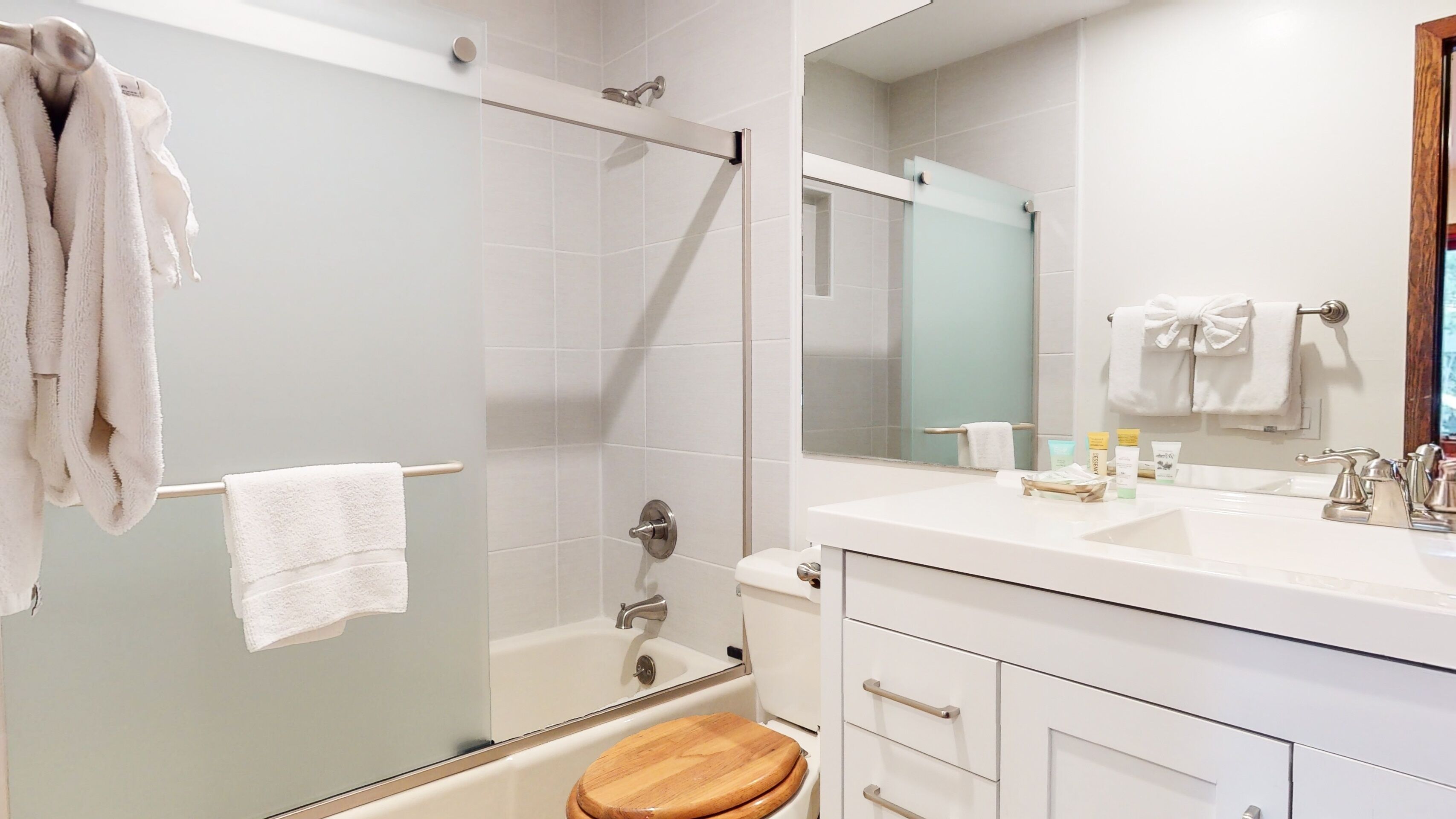 A modern bathroom with a glass shower enclosure, a wooden toilet seat, a white sink with toiletries, and a large mirror.