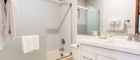 A modern bathroom with a glass shower enclosure, a wooden toilet seat, a white sink with toiletries, and a large mirror.