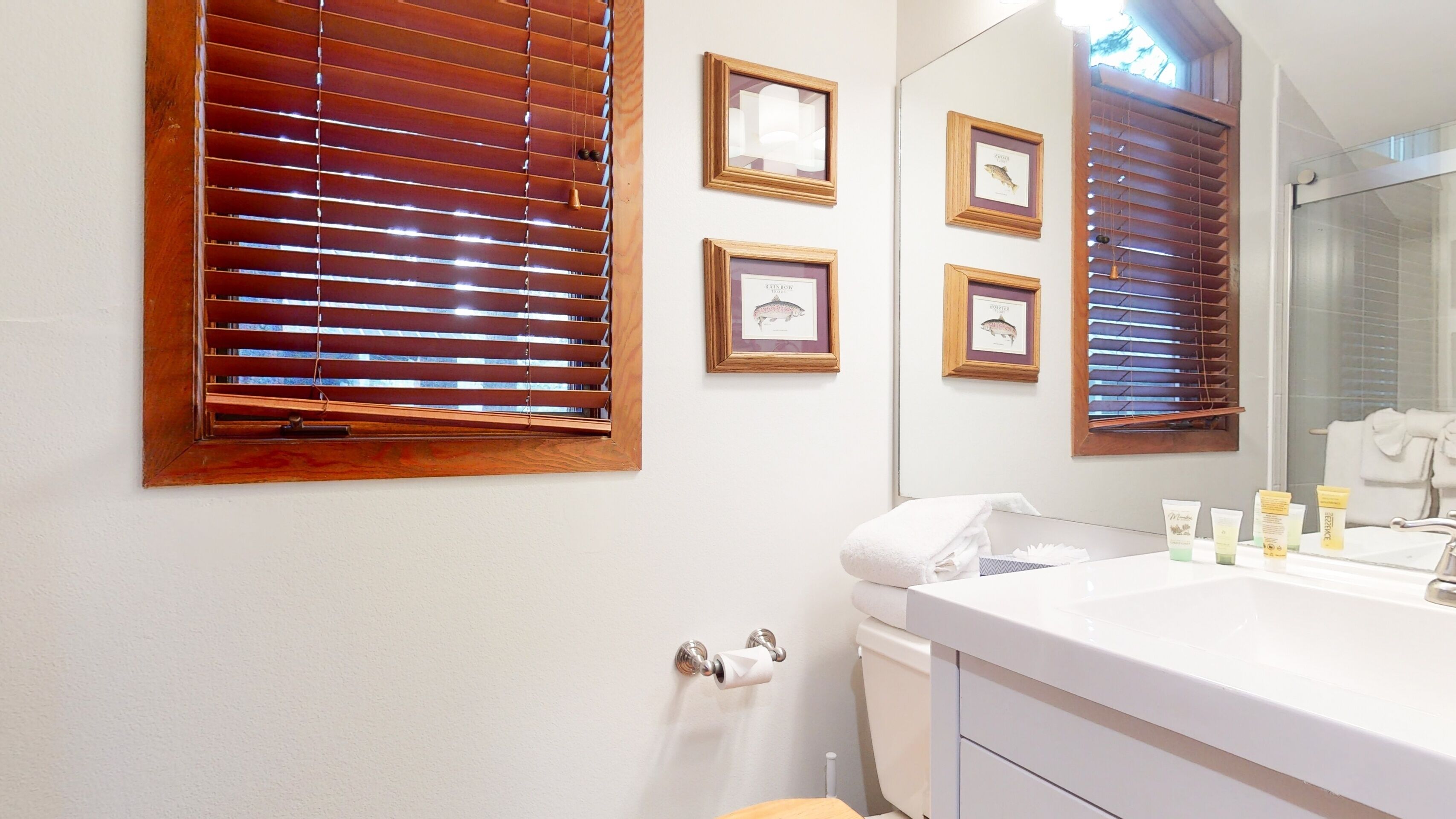 A bathroom with wooden blinds, a sink with toiletries, and framed pictures on the wall.