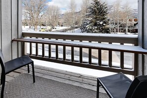 Een balkon met een houten hekwerk, twee zwarte stoelen en uitzicht op een besneeuwde straat met gebouwen en een kerstboom.