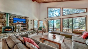 A spacious living room with a stone fireplace, a television, and large windows offering a view of trees. - Your Roomy Northstar Retreat Awaits by RedAwning (Truckee)