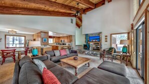 A spacious living room with a large sofa, a coffee table, and a fireplace. There is a kitchen area in the background with wooden cabinets and a dining table. - Your Roomy Northstar Retreat Awaits by RedAwning (Truckee)
