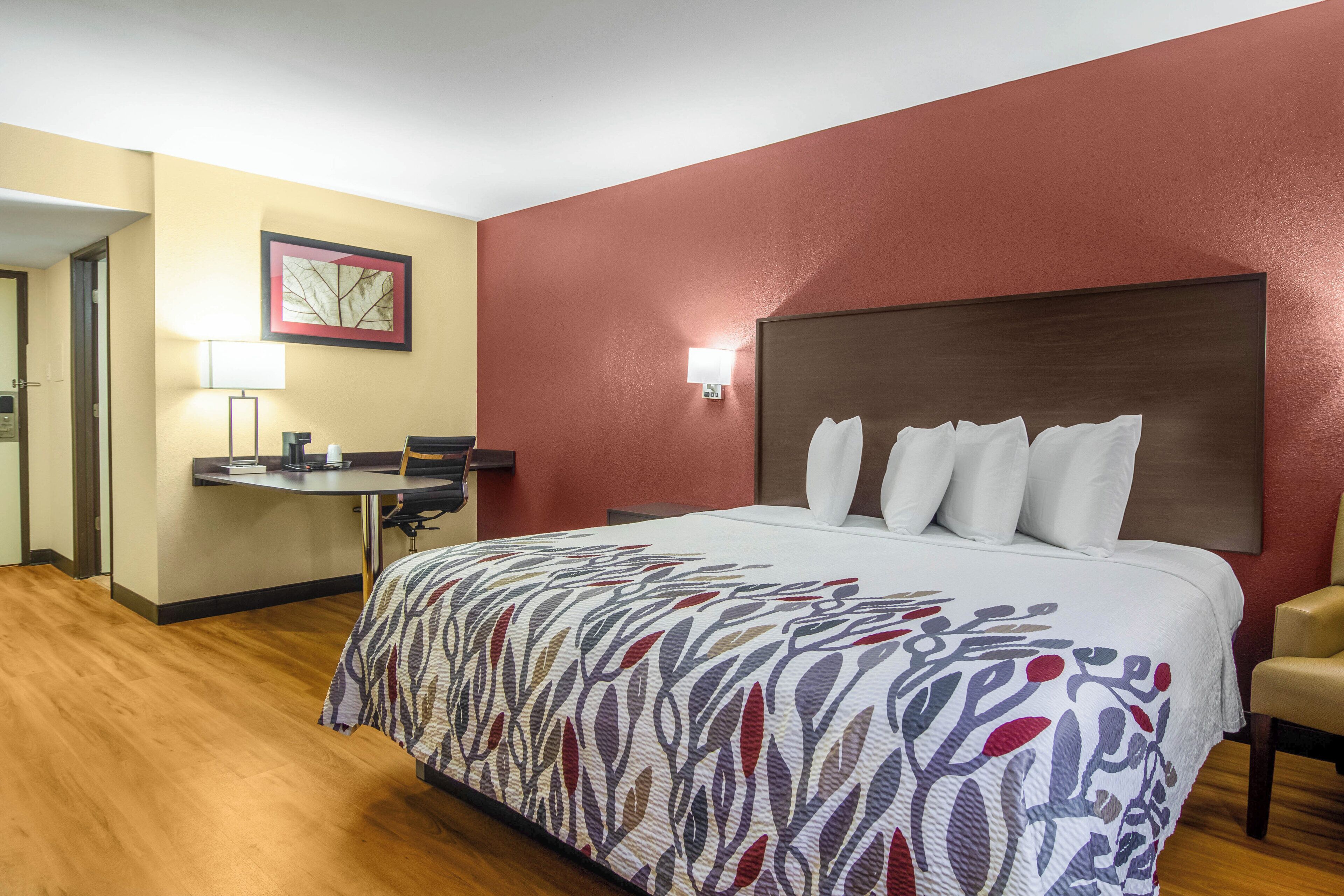 Una habitación de hotel con cama, escritorio, silla, lámpara y un cuadro en la pared.