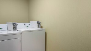 A laundry area with two washing machines and a dryer, a control panel, and a wall-mounted scale. - Red Roof Inn Perrysburg (Perrysburg)