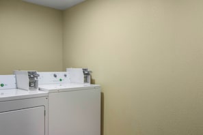 A laundry area with two washing machines and a dryer, a control panel, and a wall-mounted scale. - Red Roof Inn Perrysburg (Perrysburg)