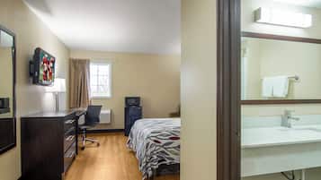 A hotel room with a bed, a desk, a chair, a television, a window with curtains, a refrigerator, and a bathroom with a sink and mirror.