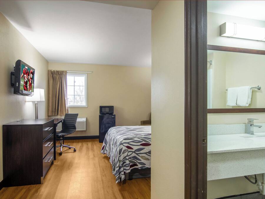 Een hotelkamer met een bed, bureau, stoel, televisie, raam met gordijnen, koelkast en een badkamer met wastafel en spiegel.