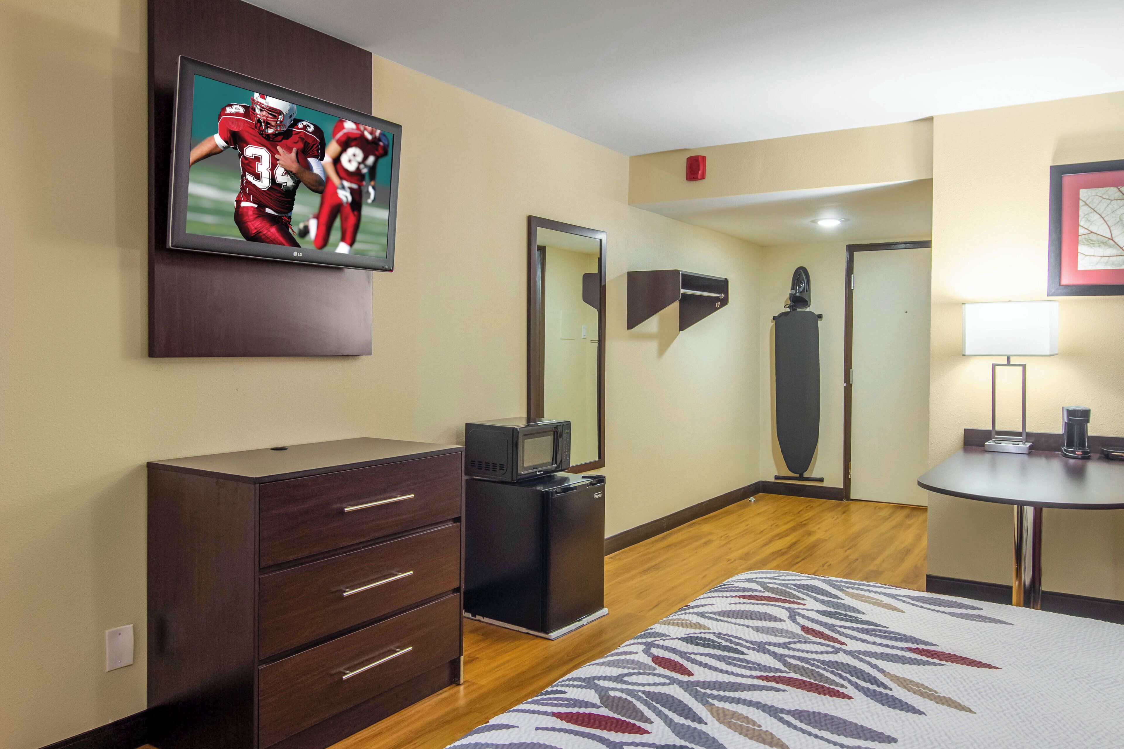 A hotel room with a flat-screen TV mounted on the wall, a dresser with drawers, a microwave, a bed with a patterned bedspread, and a small table with a lamp.