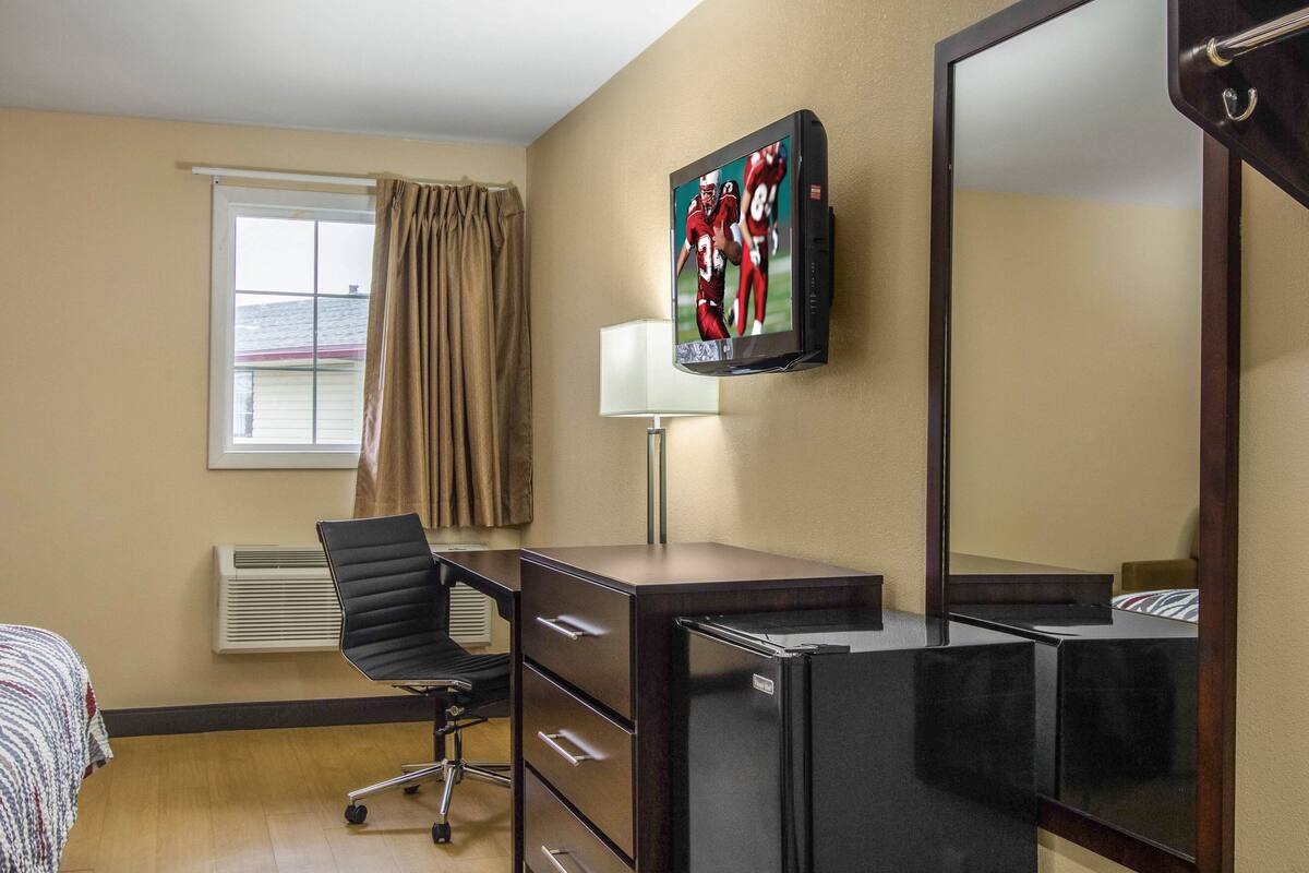 Quarto de hotel com cama, escrivaninha, cadeira, televisão fixada na parede e janela com cortinas.