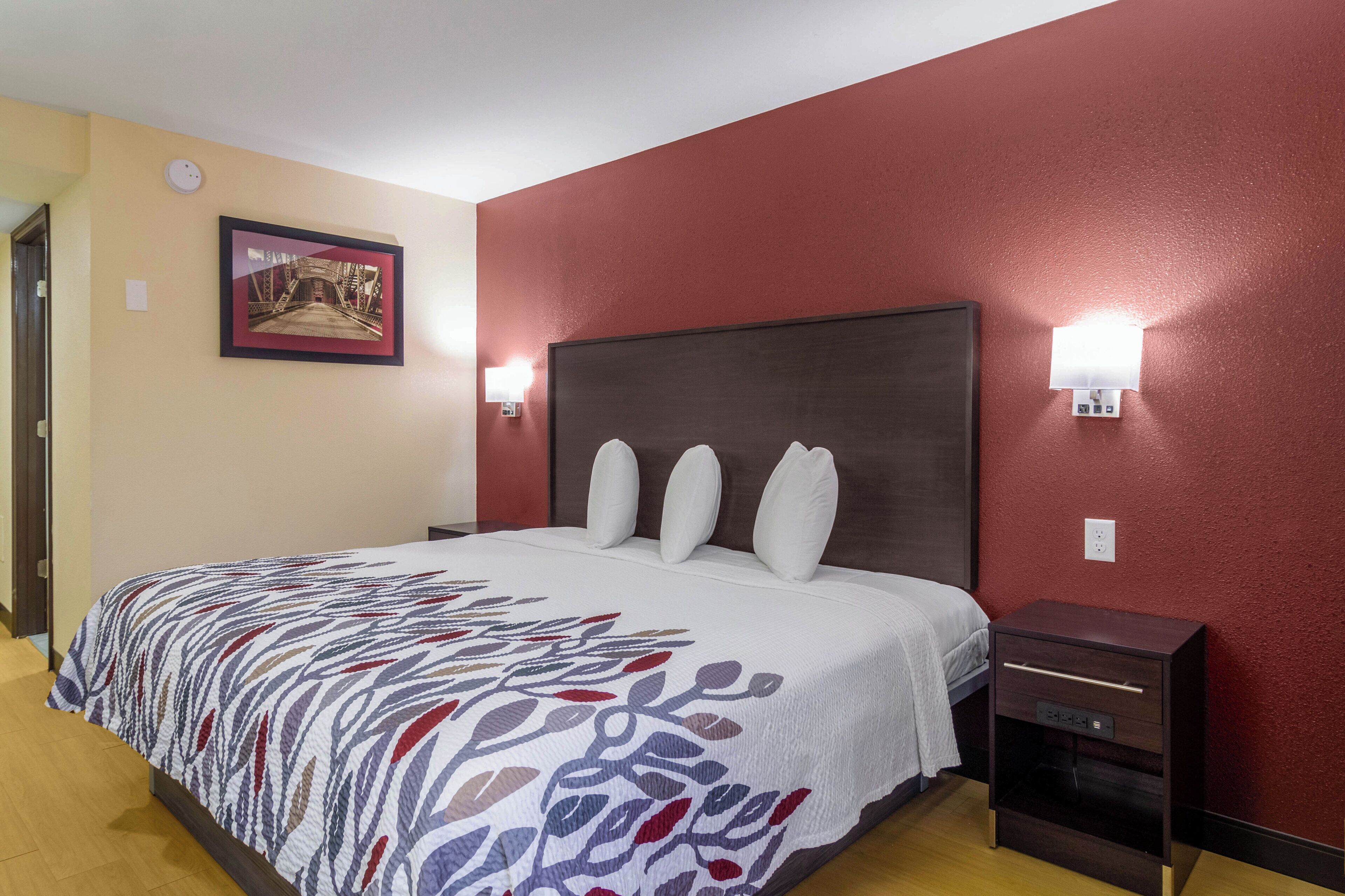 A hotel room with a bed, a nightstand, a wall-mounted lamp, and a framed picture on the wall.