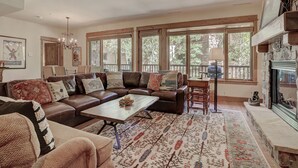 A spacious living room with a leather sofa, a coffee table, and a patterned rug. There are large windows with blinds, a chandelier, and a fireplace. - Elegant Updated 3 Bedroom Condo At One Arrowhead Place Condo by RedAwning (Edwards)