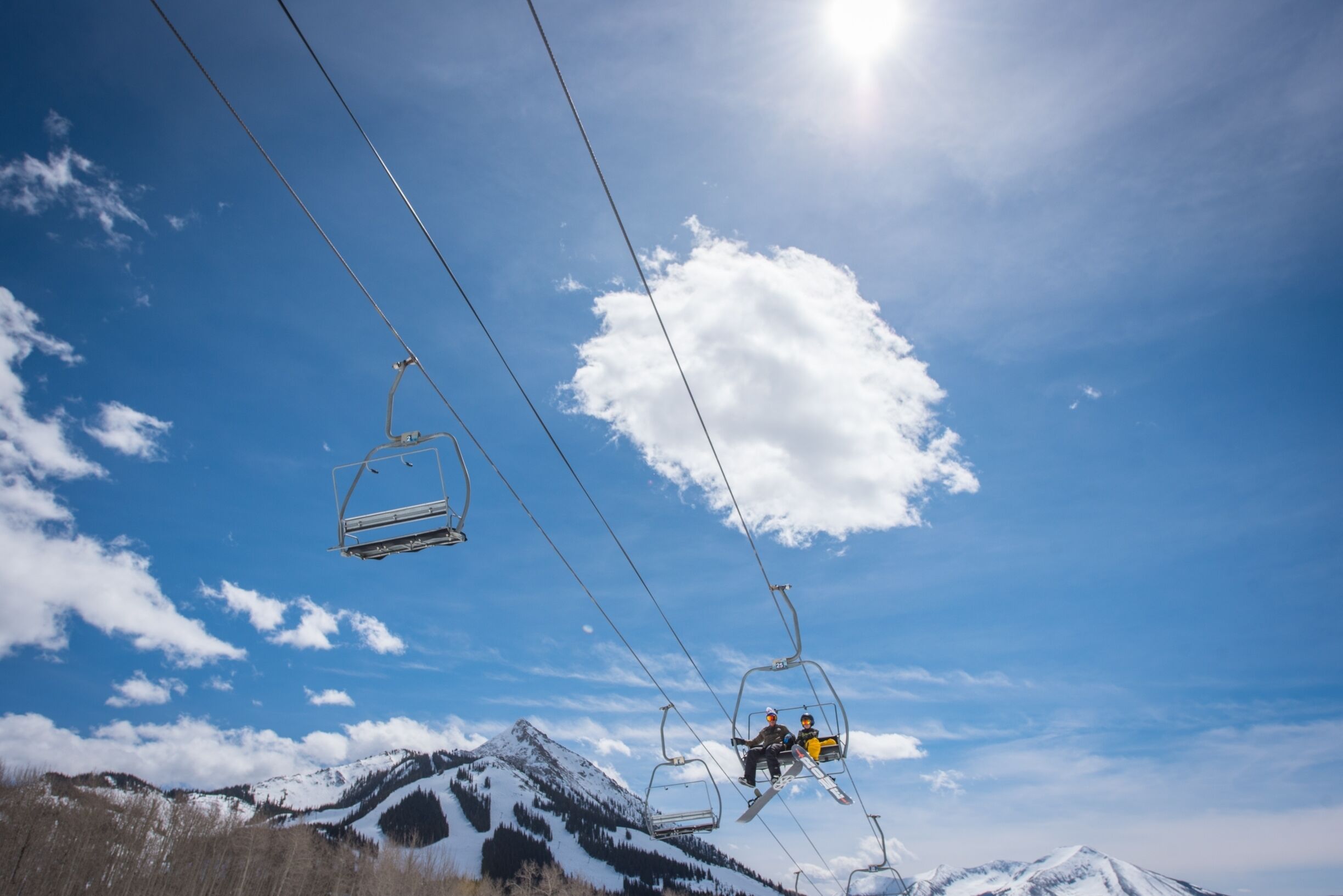 Eine Sesselbahn mit zwei Sesseln, die Fahrgäste befördert, vor einer Kulisse aus schneebedeckten Bergen und blauem Himmel.