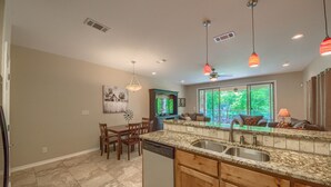 A modern kitchen with granite countertops, wooden cabinets, and a dishwasher. There is a dining area with a table and chairs, and a large window providing a view of greenery outside. - Riverview Terrace Rg 203 3 Bedroom Condo by RedAwning (New Braunfels)
