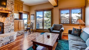 A cozy living room with a stone fireplace, wooden beams, a large window with a mountain view, a comfortable sofa, and a coffee table. - Unique & Modern 3 Br/2ba - Ski-in/ski-out 3 Bedroom Condo - No Cleaning Fee! by RedAwning (Crested Butte)