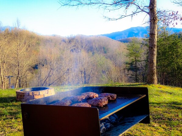 A grill with meat being cooked outdoors, surrounded by trees and a hilly landscape.