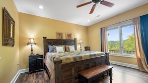 A bedroom with a wooden bed, two bedside tables, a bench, a ceiling fan, and a large window with a view. - Enchanted Primrose 2 Bedroom Chalet by RedAwning (Sevierville)