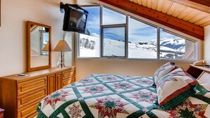 A bedroom with a large window offering a mountain view, a wooden dresser, a television, and a quilted bedspread. - 1 Br + Loft With Mountain Views 1 Bedroom Condo - No Cleaning Fee! by RedAwning (Crested Butte)