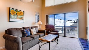 A living room with a brown sofa, a coffee table, a lamp, and a large window with a view of a snowy landscape. - 1 Br + Loft With Mountain Views 1 Bedroom Condo - No Cleaning Fee! by RedAwning (Crested Butte)